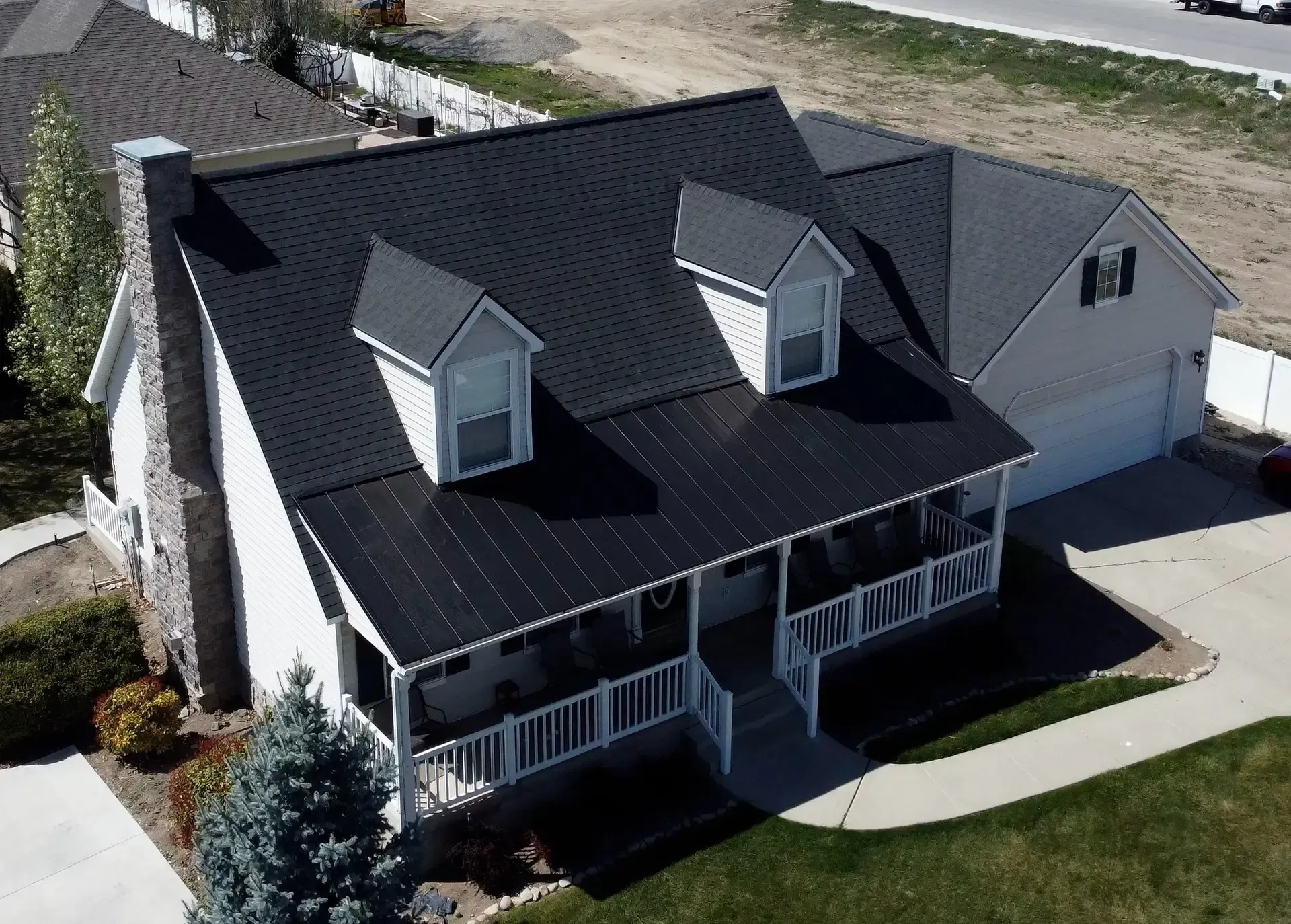 Black metal roof on a white house with a porch, two dormers, and a stone chimney, aerial view.