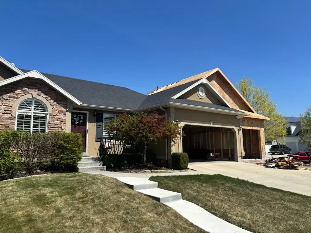 House with open garage and roof construction against a blue sky. Green lawn and shrubs in the foreground.