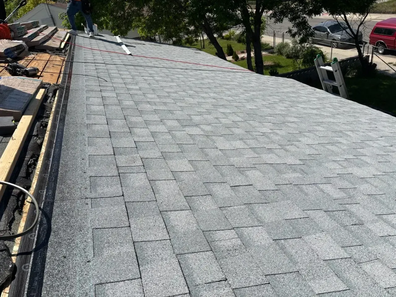 Gray asphalt shingles on a roof, under construction.  A ladder, cars, and trees are in the background.