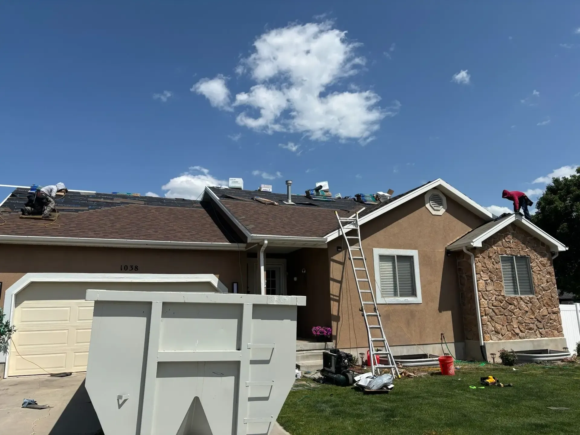 Roofers replacing shingles on a house under a partly cloudy blue sky; a dumpster and ladder are in the yard.