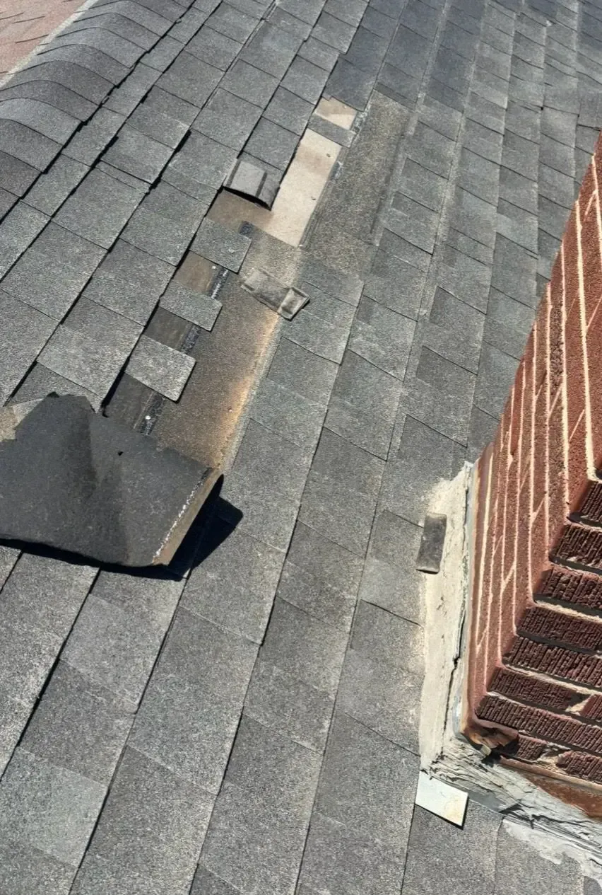 Damaged asphalt shingles on a roof near a brick chimney. Shingles are missing or broken, exposing the underlayment.
