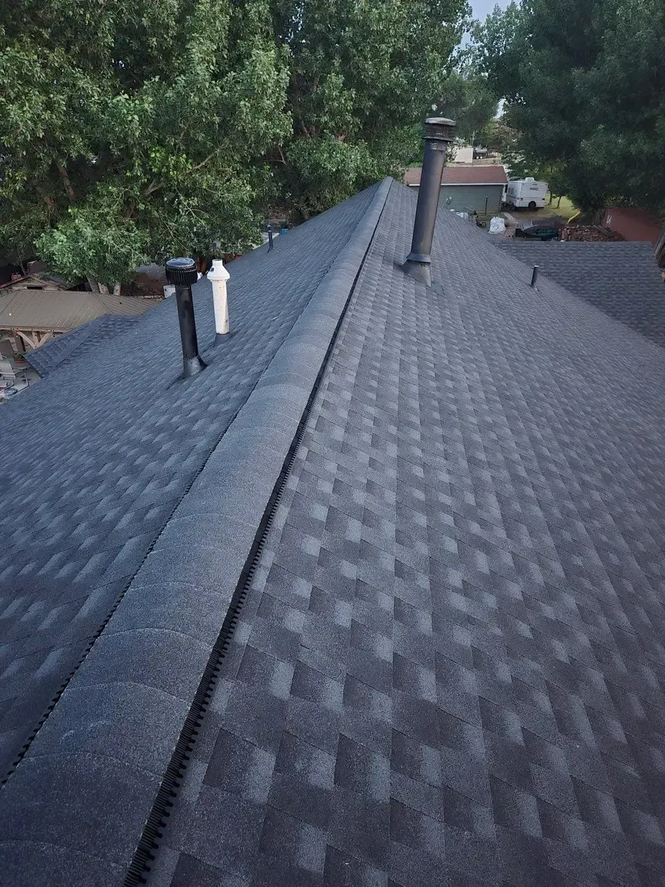 Dark gray asphalt shingle roof with two vents and a chimney against a background of trees.