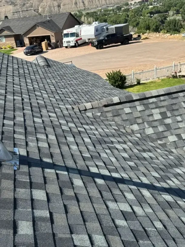 A close-up view of a shingled roof with a background of houses, vehicles, and a hillside.
