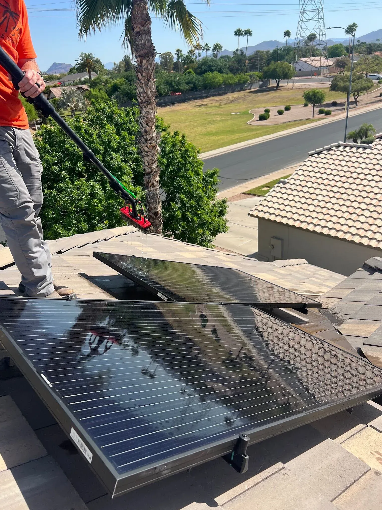 Person cleaning solar panels on a rooftop with a long-handled brush, sunny day.