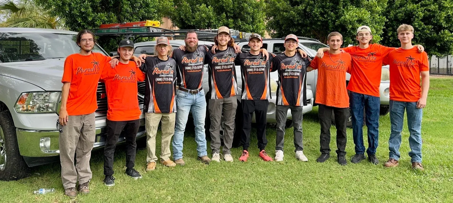 A group of ten people stand on grass, some wearing orange shirts, some black shirts, posing in front of two silver trucks.