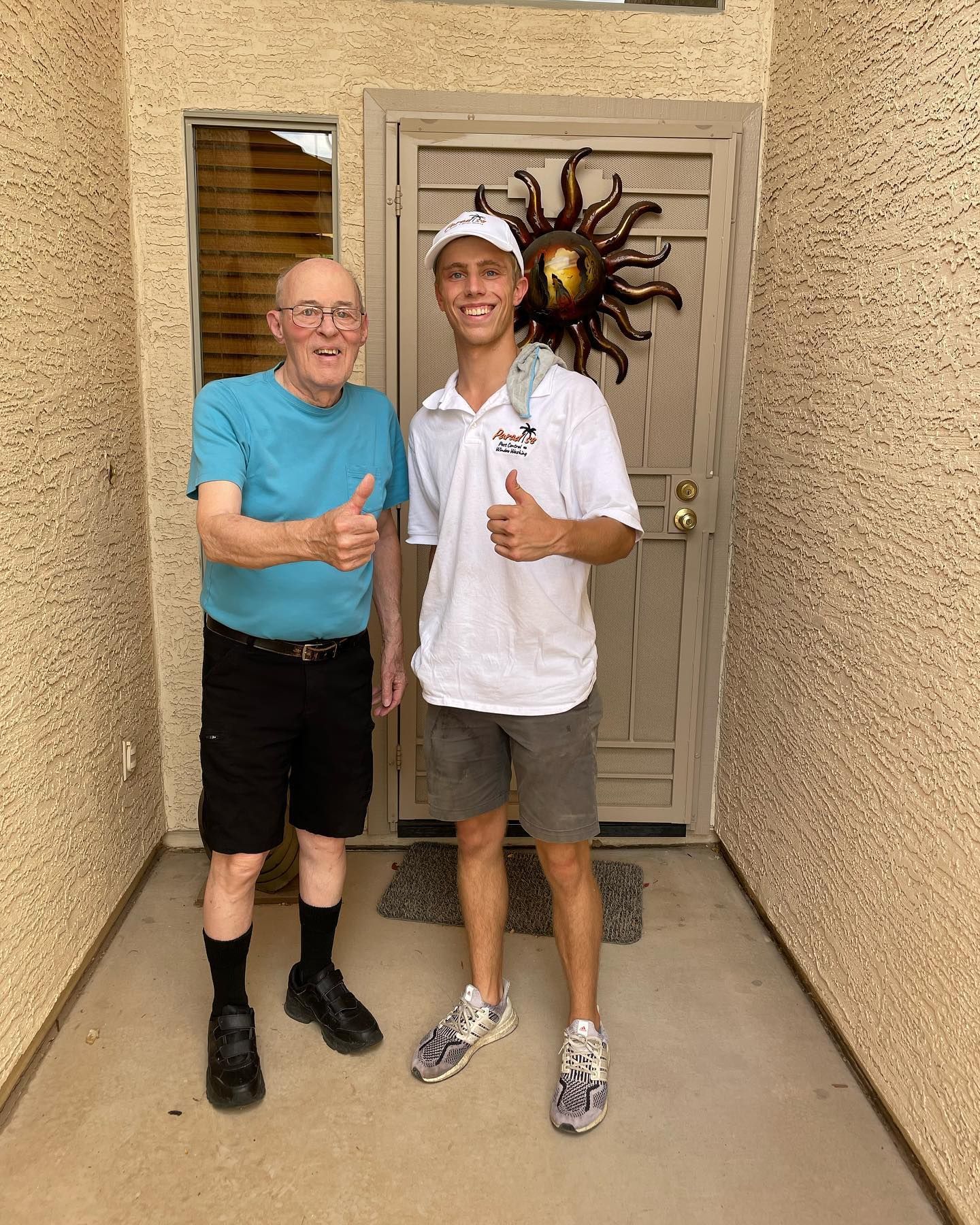 Two men are standing next to each other in front of a door giving a thumbs up.
