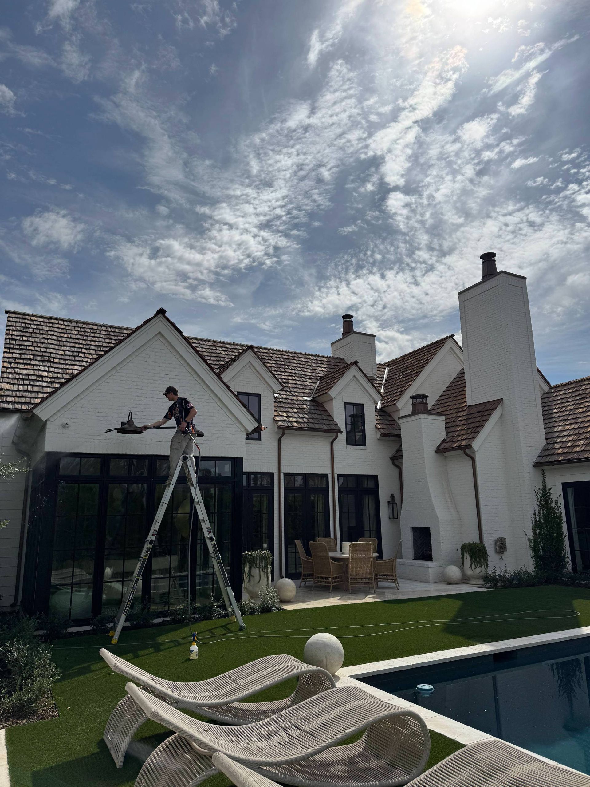 Person on a ladder painting a white house with brown roof. Pool and sunny sky in the background.