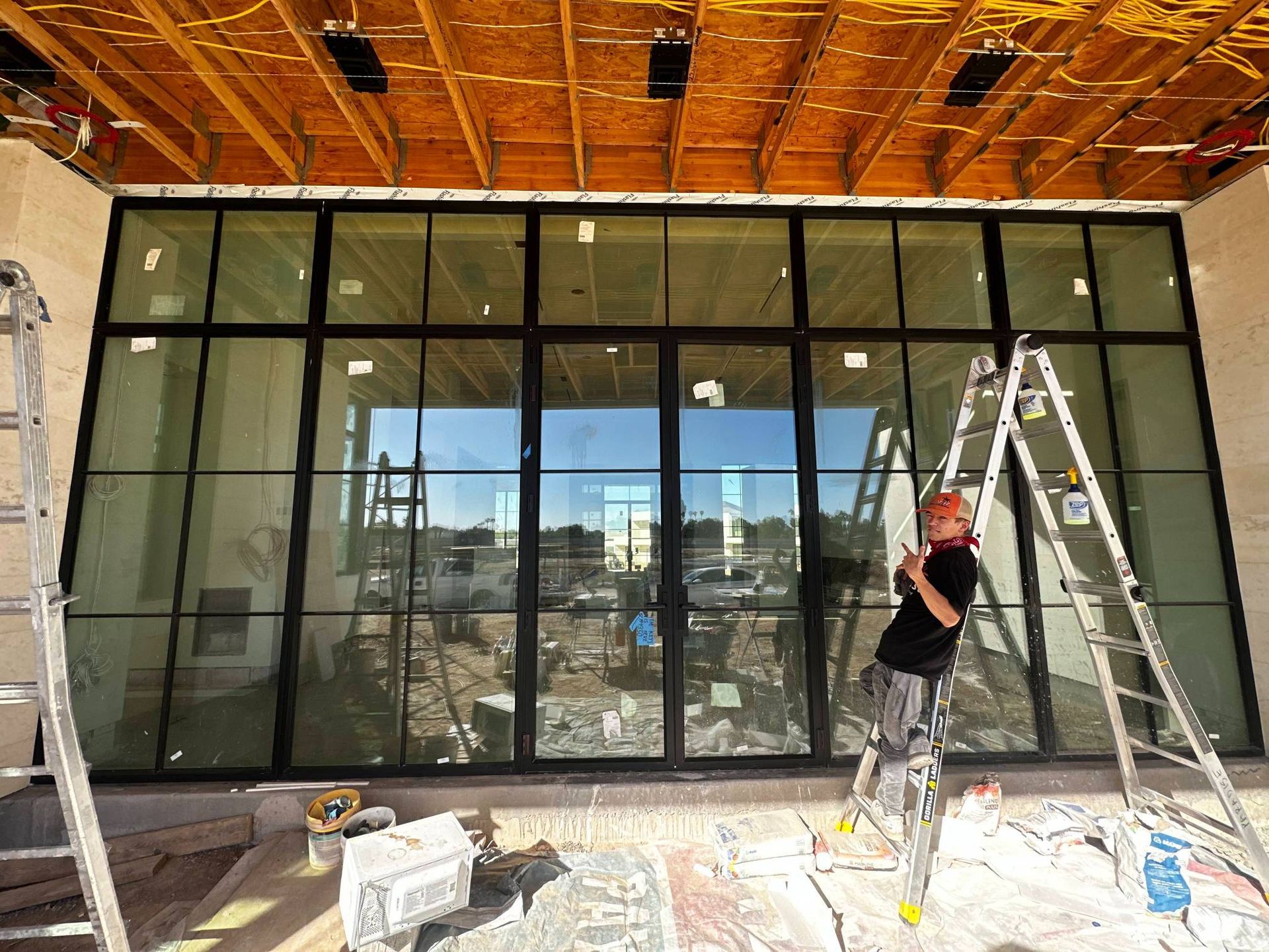 Construction worker on a ladder installing a large black-framed window. Building in progress. Sunny setting.