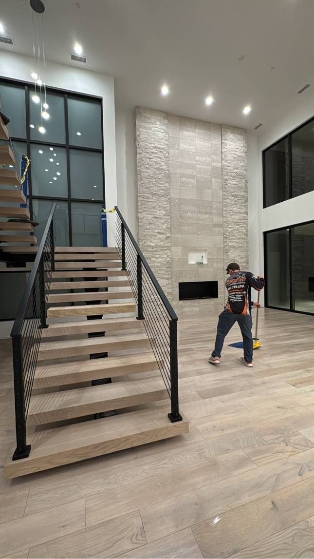 Person sweeping wooden floor of a large room with staircase and fireplace.