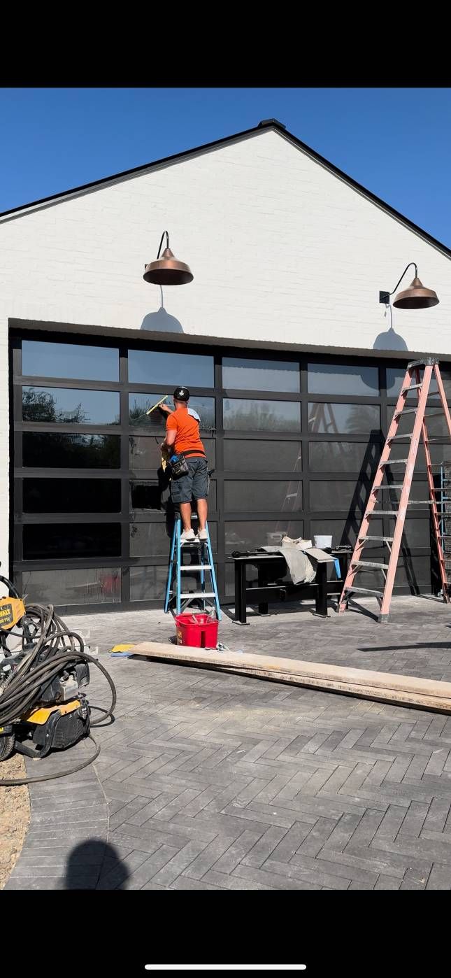 A person on a ladder cleans a glass garage door. Construction tools and a second ladder are nearby.