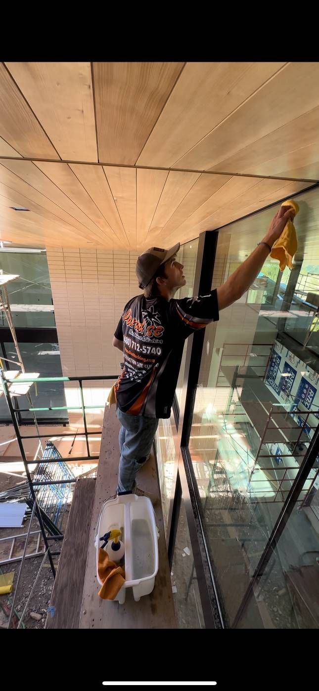 Person cleaning a large window with a yellow cloth. They stand on a ledge with a bucket below.