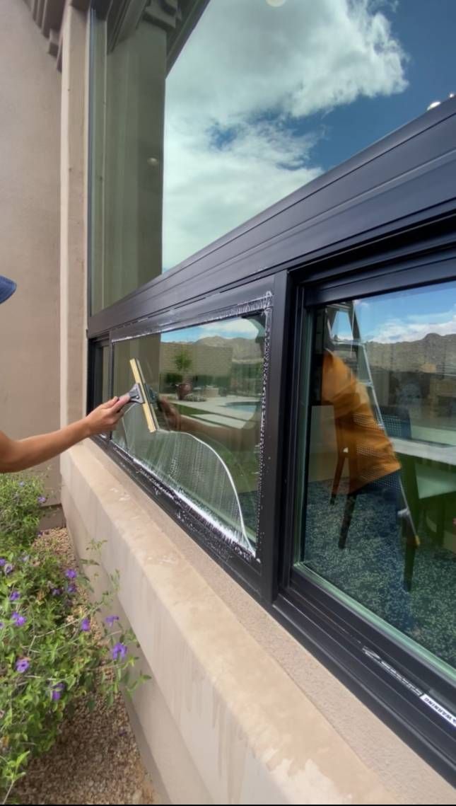 Person using a squeegee to clean a large window on a beige building with a blue sky background.
