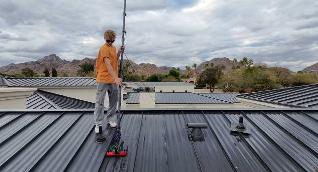 Person on a roof using a cleaning tool. Cloudy sky, mountains in the background, gray metal roof.