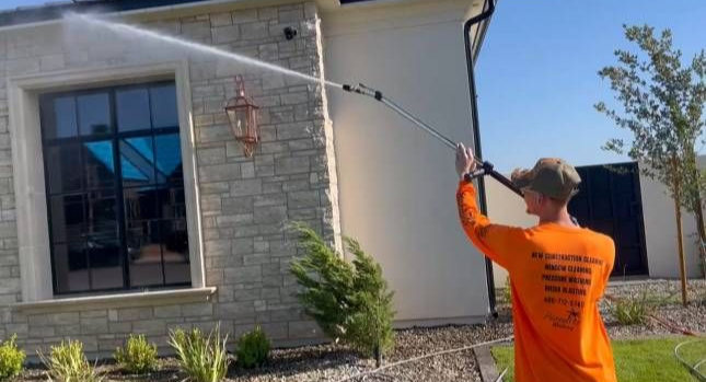 Man in orange shirt pressure washing a stone house exterior.