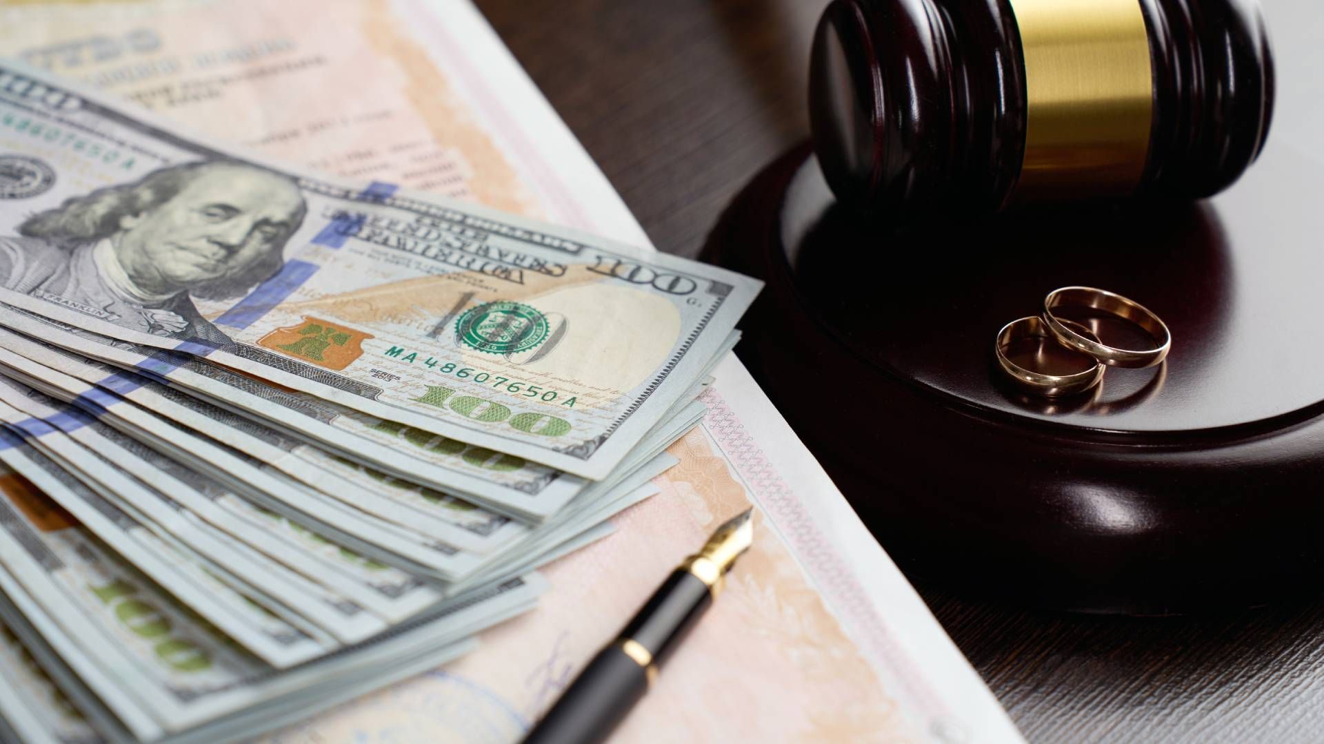 A stack of money , a pair of wedding rings , a pen , and a gavel on a table.