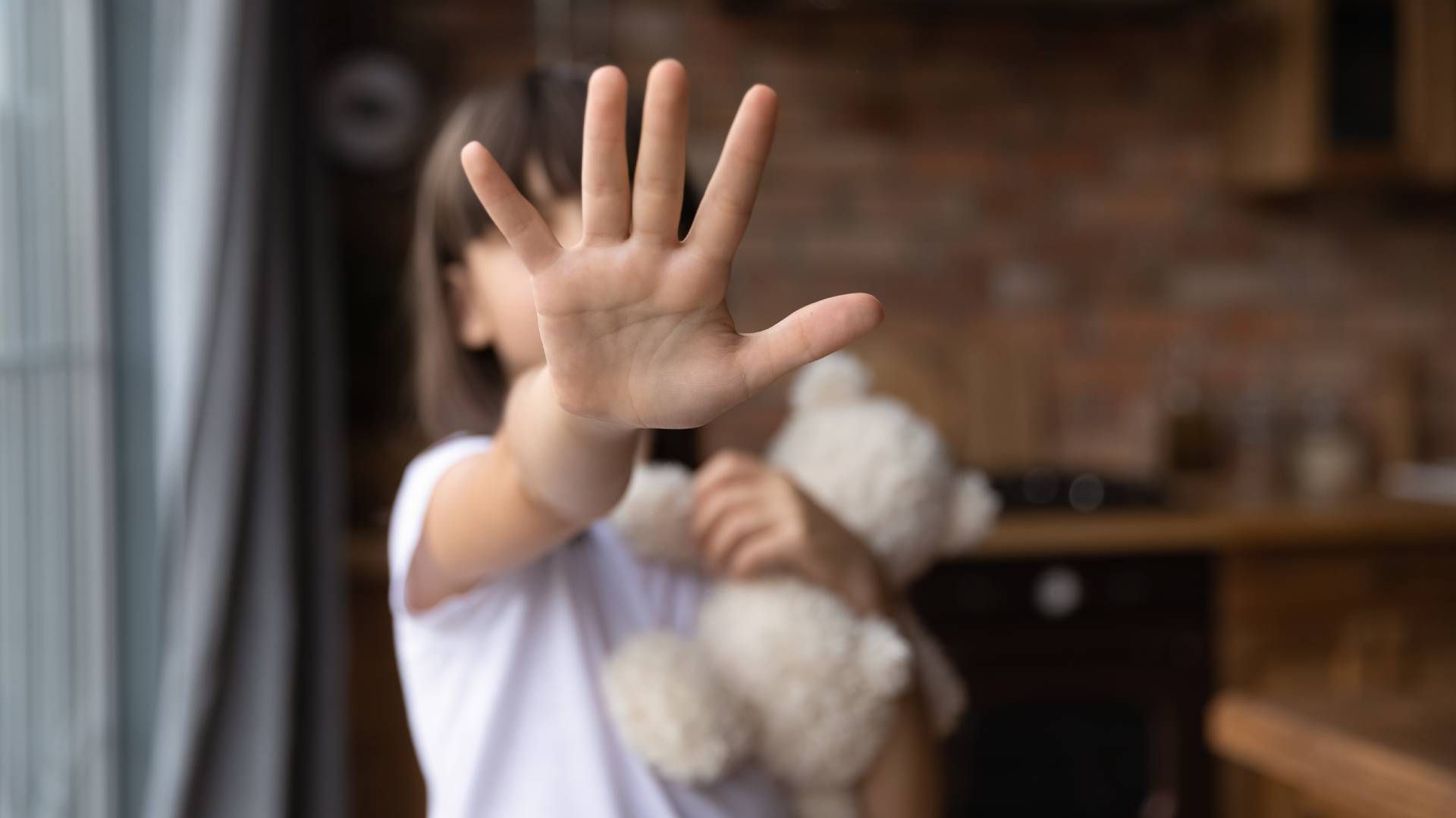 A little girl is holding a teddy bear and making a stop sign with her hand.