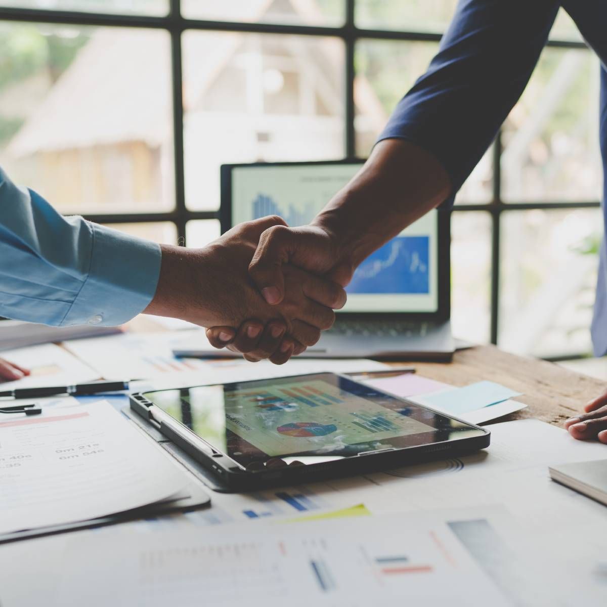 Two people shaking hands over a table with a laptop and tablet