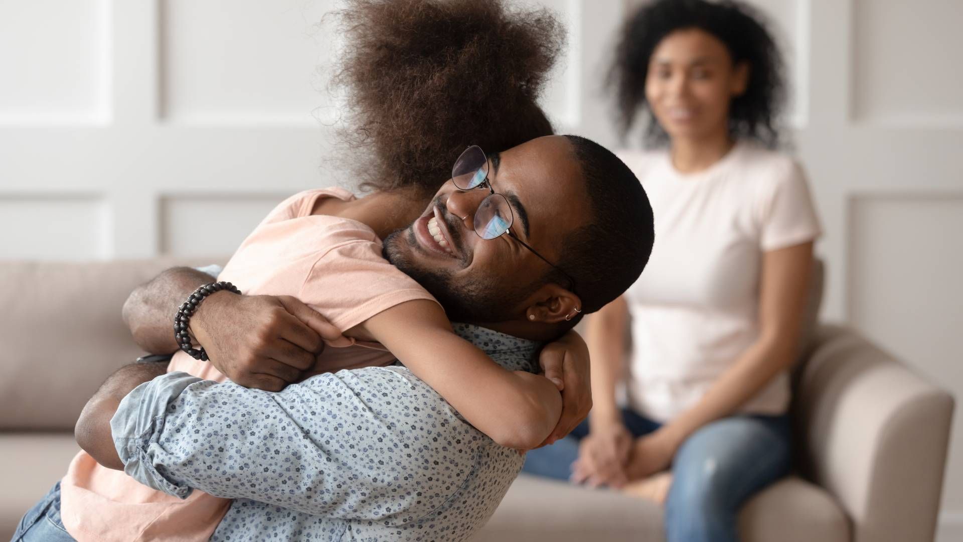 A man is hugging a little girl while a woman sits on a couch.
