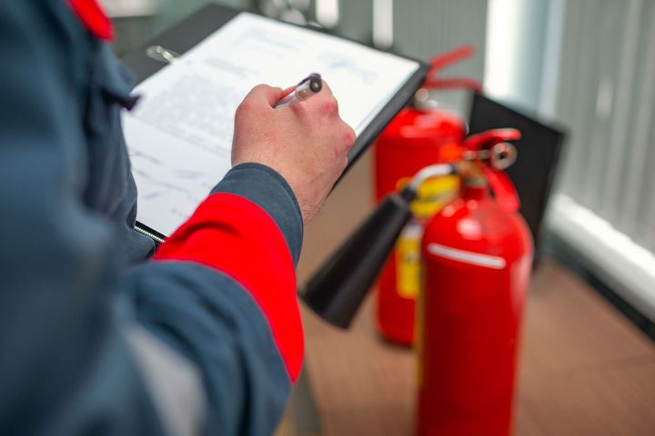 A man is writing on a clipboard next to fire extinguishers.