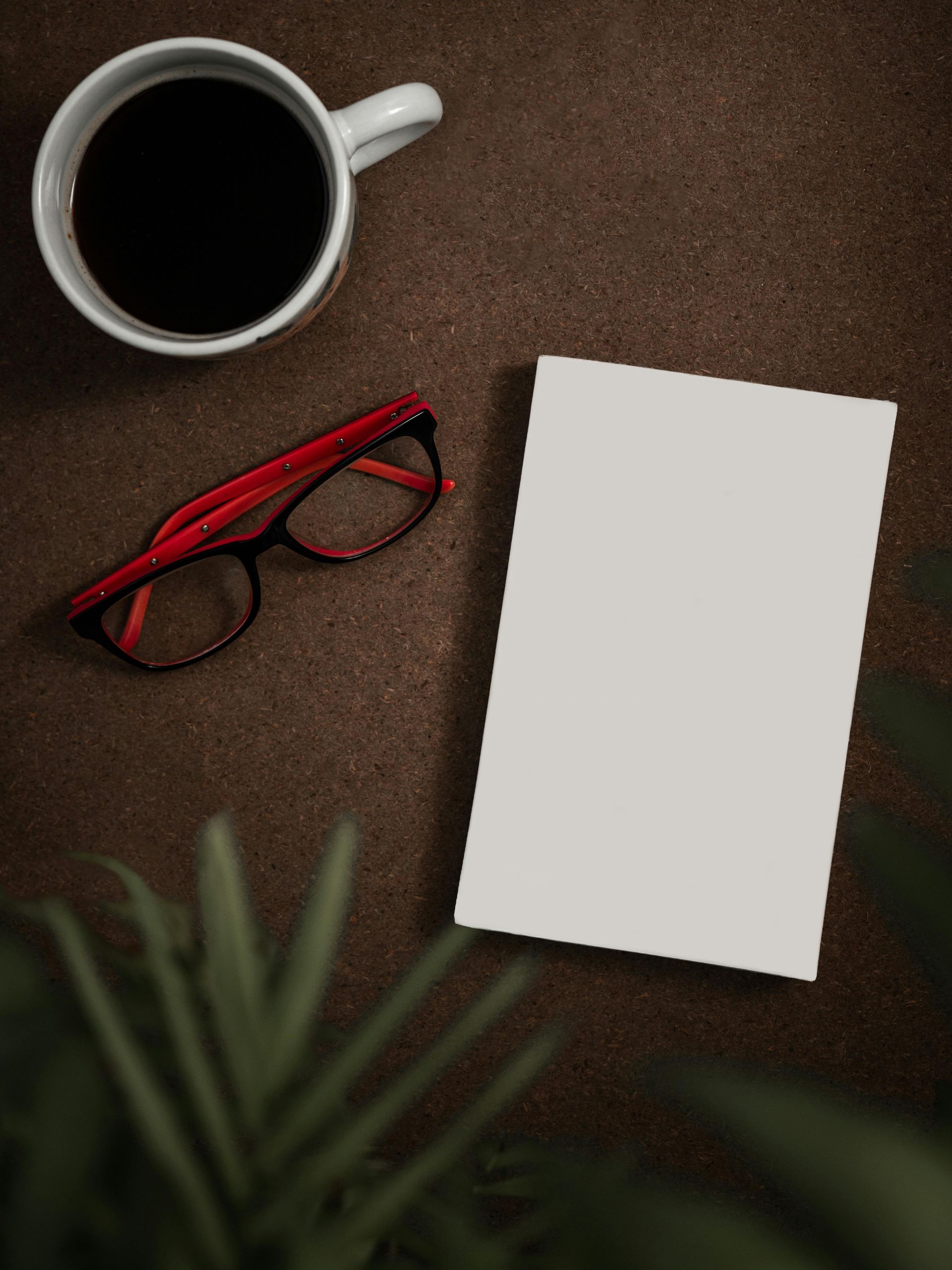A cup of coffee , a book , glasses and a plant on a table.