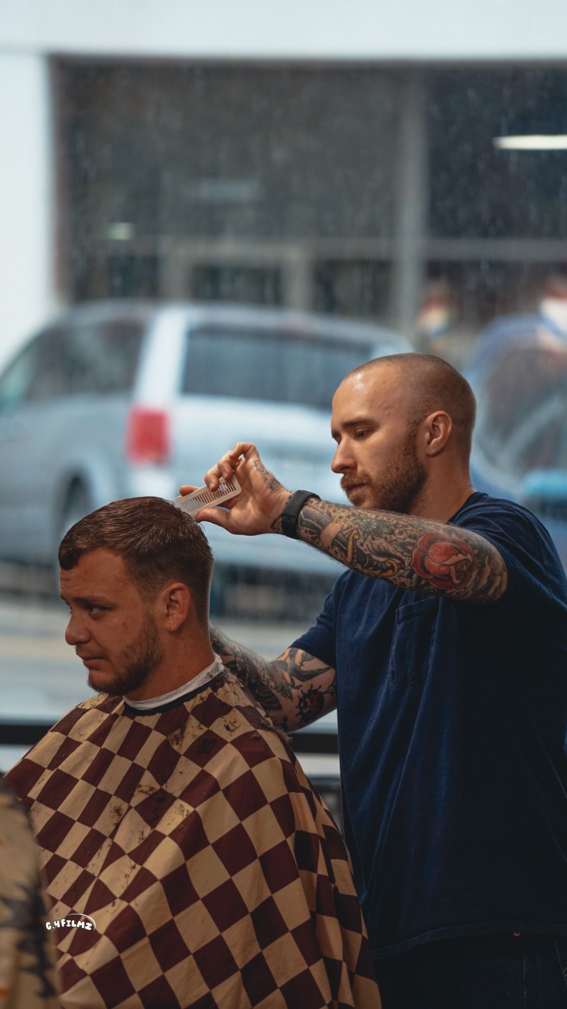 Barber cutting a client's hair in a shop, both wearing capes. Rain falls outside the window.