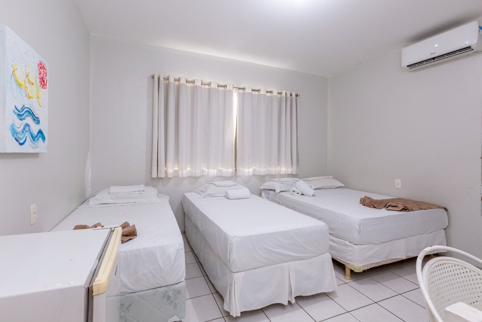 Um quarto de hotel com três camas de solteiro com lençóis brancos, paredes brancas, uma janela com cortinas e ar condicionado.