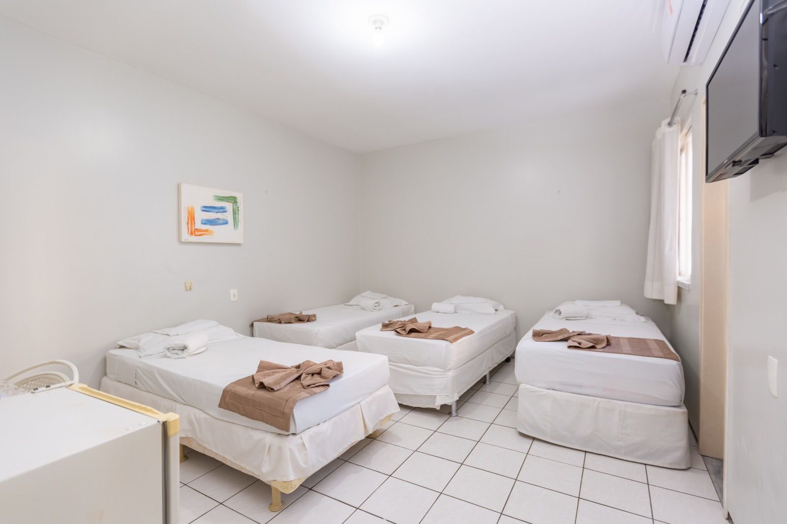 Um quarto de hotel com paredes brancas, piso de azulejo e três camas de solteiro com lençóis brancos e cobertores bege.