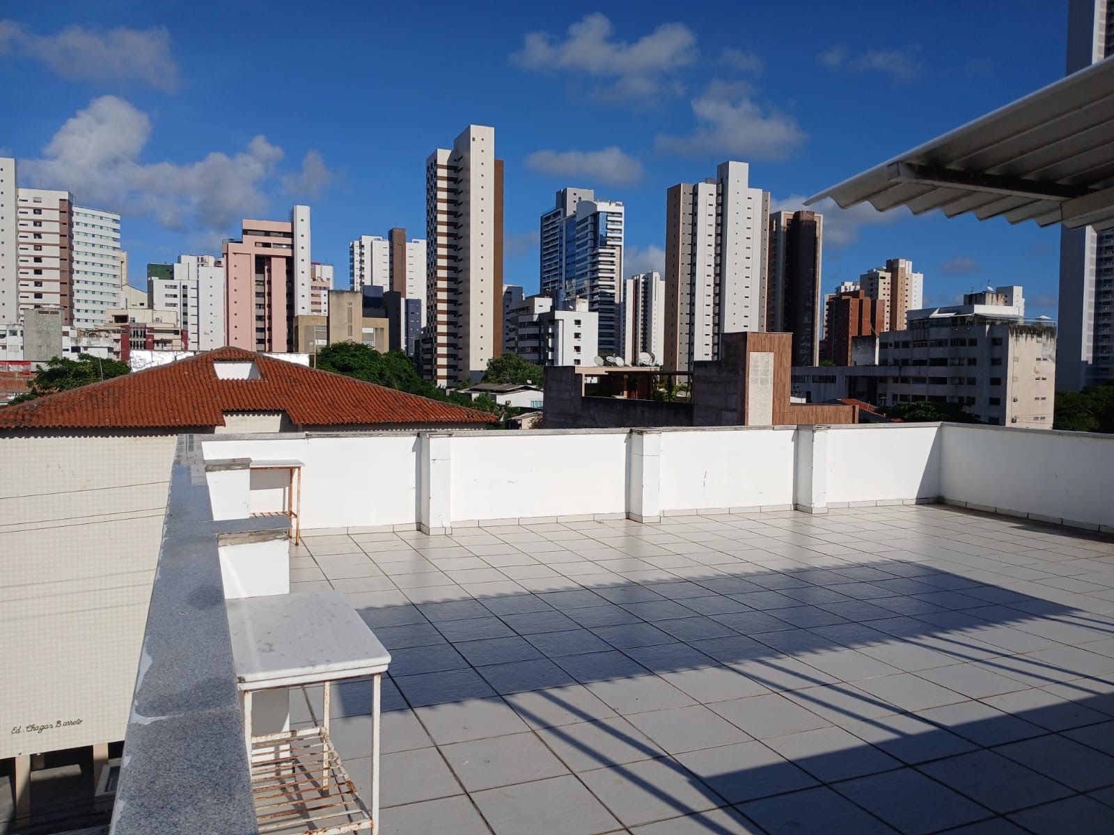 Um terraço na cobertura com azulejos claros, com vista para uma paisagem urbana de prédios residenciais altos sob um céu azul límpido.