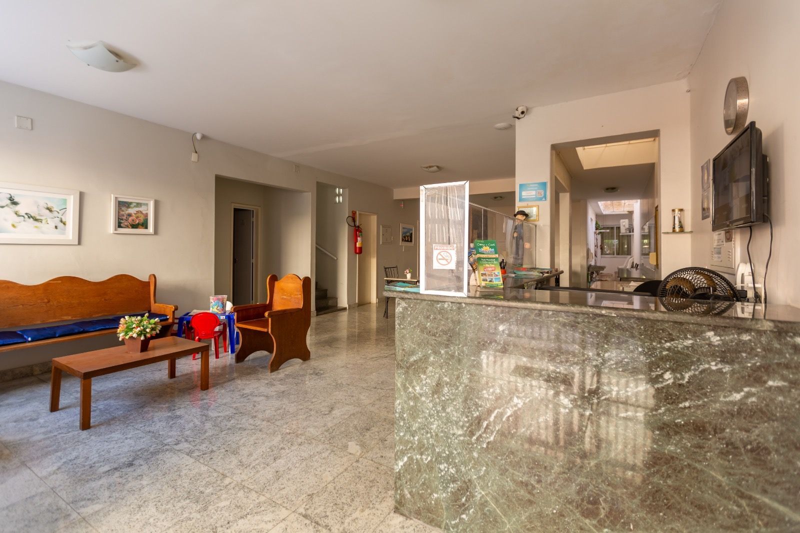 Uma área de recepção com uma mesa de mármore verde, um banco de madeira, uma pequena mesa com uma planta e uma poltrona marrom sobre um piso de azulejos.