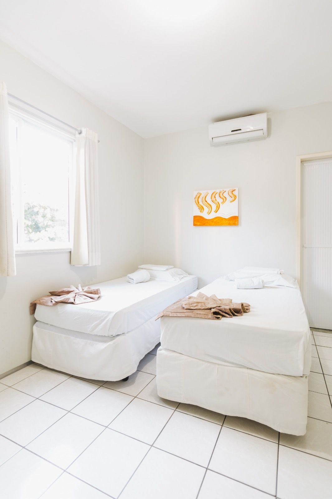 Um quarto luminoso e minimalista com duas camas de solteiro com lençóis brancos, piso de azulejo e um pequeno quadro em uma parede branca.