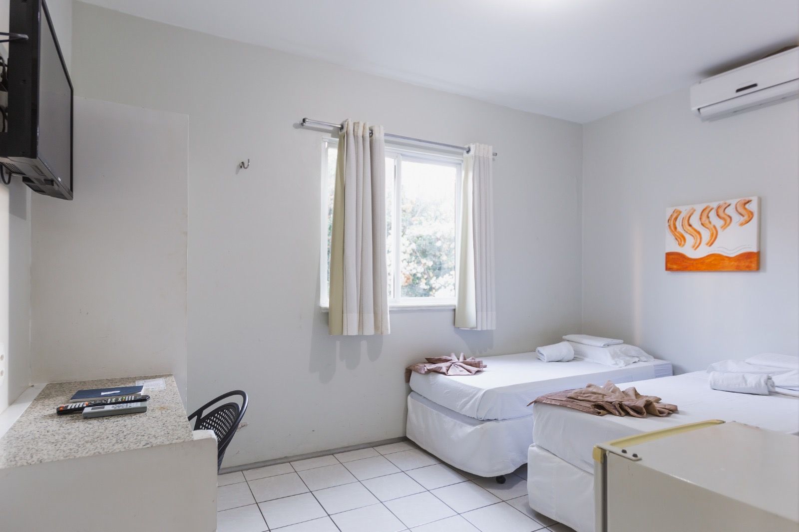 Um quarto com duas camas de solteiro, paredes brancas, piso de azulejo branco, uma escrivaninha, uma televisão e uma pintura abstrata.