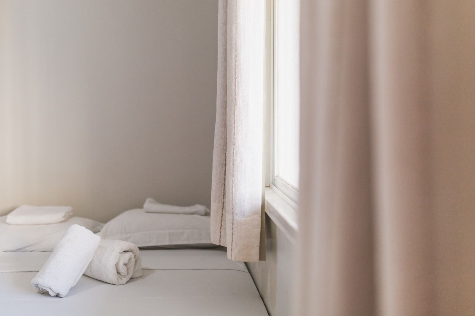 Duas toalhas brancas enroladas estão sobre uma cama com travesseiros, ao lado de uma janela com cortinas bege-claras em um quarto minimalista.
