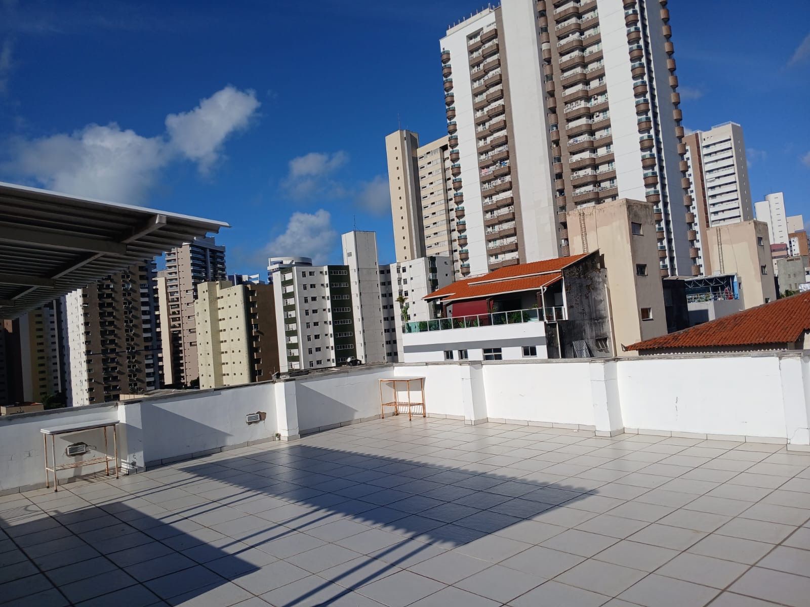 Um terraço na cobertura com piso de azulejos, com vista para o horizonte da cidade, com vários prédios altos de apartamentos sob um céu azul e nublado.