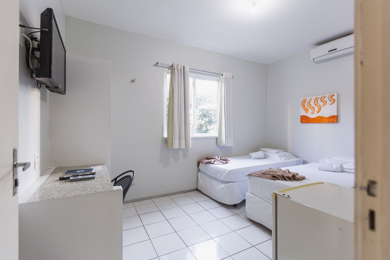Um quarto de hotel luminoso e simples, com paredes brancas, piso de azulejo, duas camas de solteiro, uma TV fixada na parede e ar-condicionado.