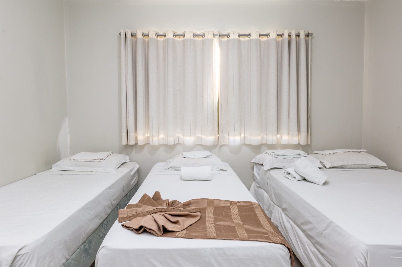 Um quarto de hotel limpo e luminoso, com três camas de solteiro com lençóis brancos, paredes brancas e cortinas translúcidas na janela.