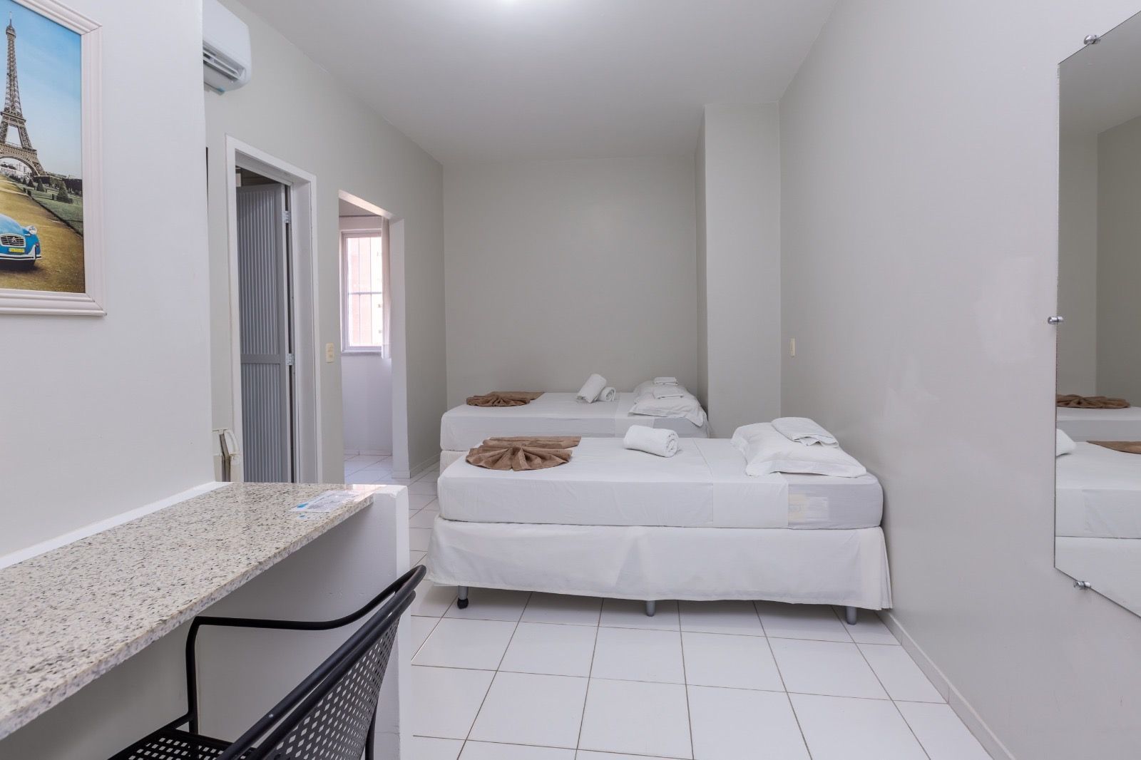 Um quarto de hotel com duas camas brancas, uma escrivaninha com uma cadeira preta, piso de azulejo e uma gravura da Torre Eiffel na parede.