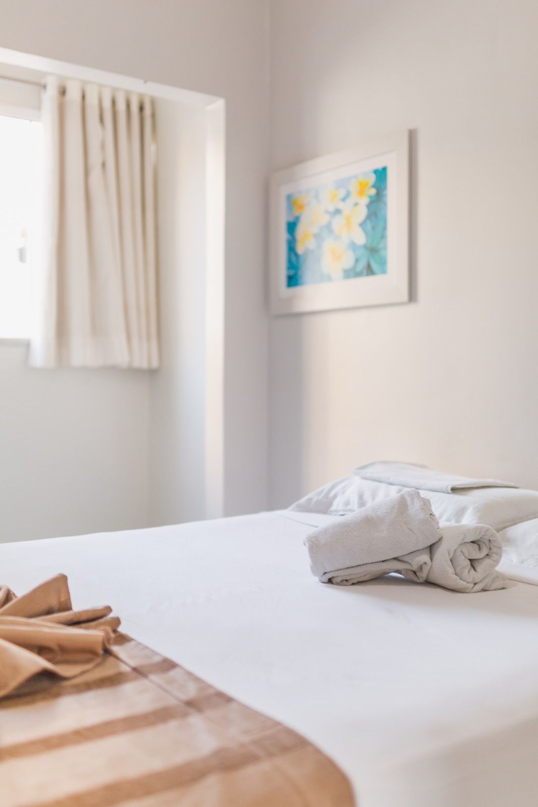 Um quarto de hotel com paredes brancas, uma cama com toalhas brancas dobradas e um caminho de mesa bege, com uma gravura floral emoldurada na parede.