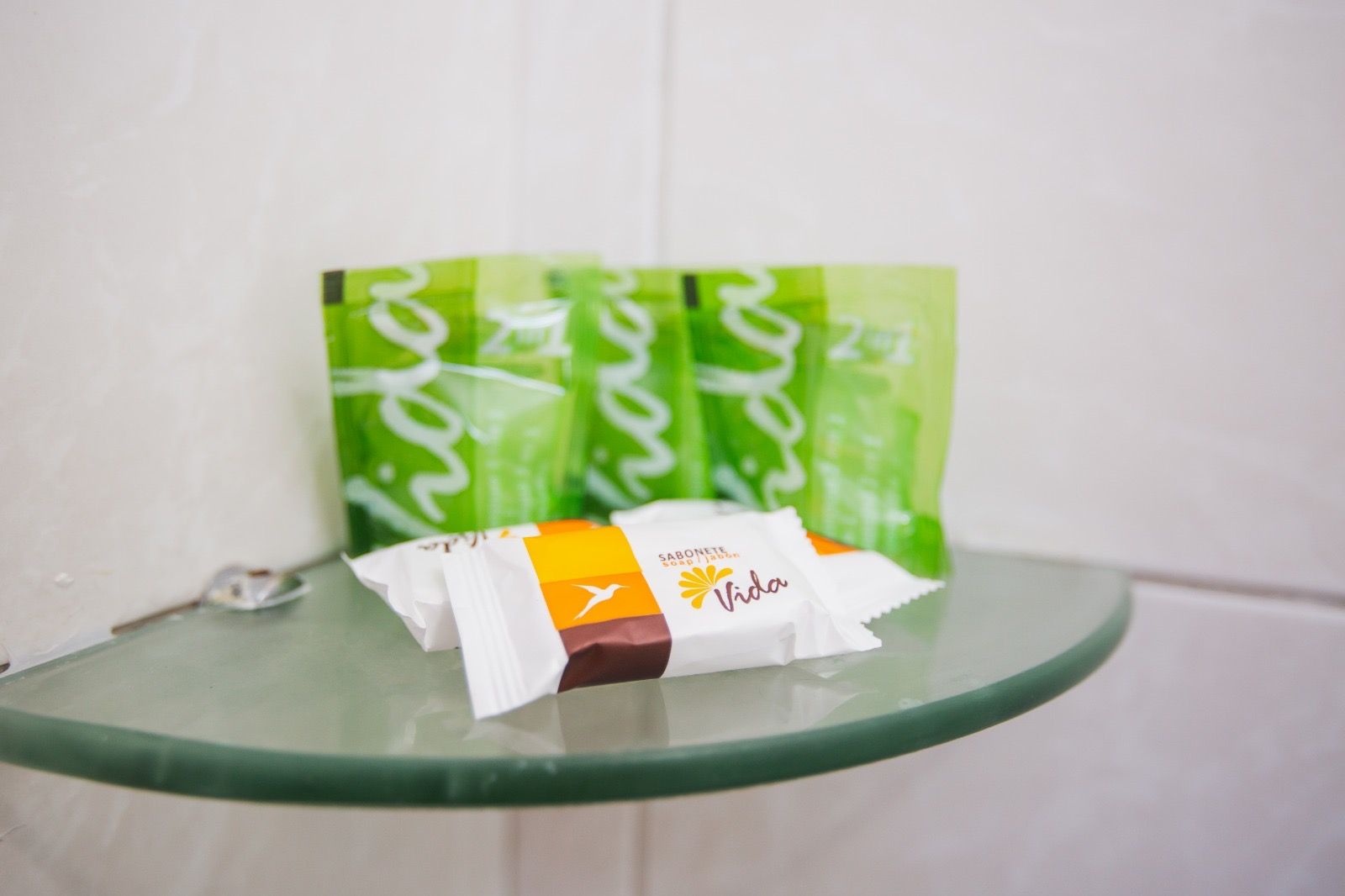 Artigos de higiene do hotel, incluindo pequenos pacotes verdes e barras de sabonete embaladas individualmente, em uma prateleira de vidro no banheiro.