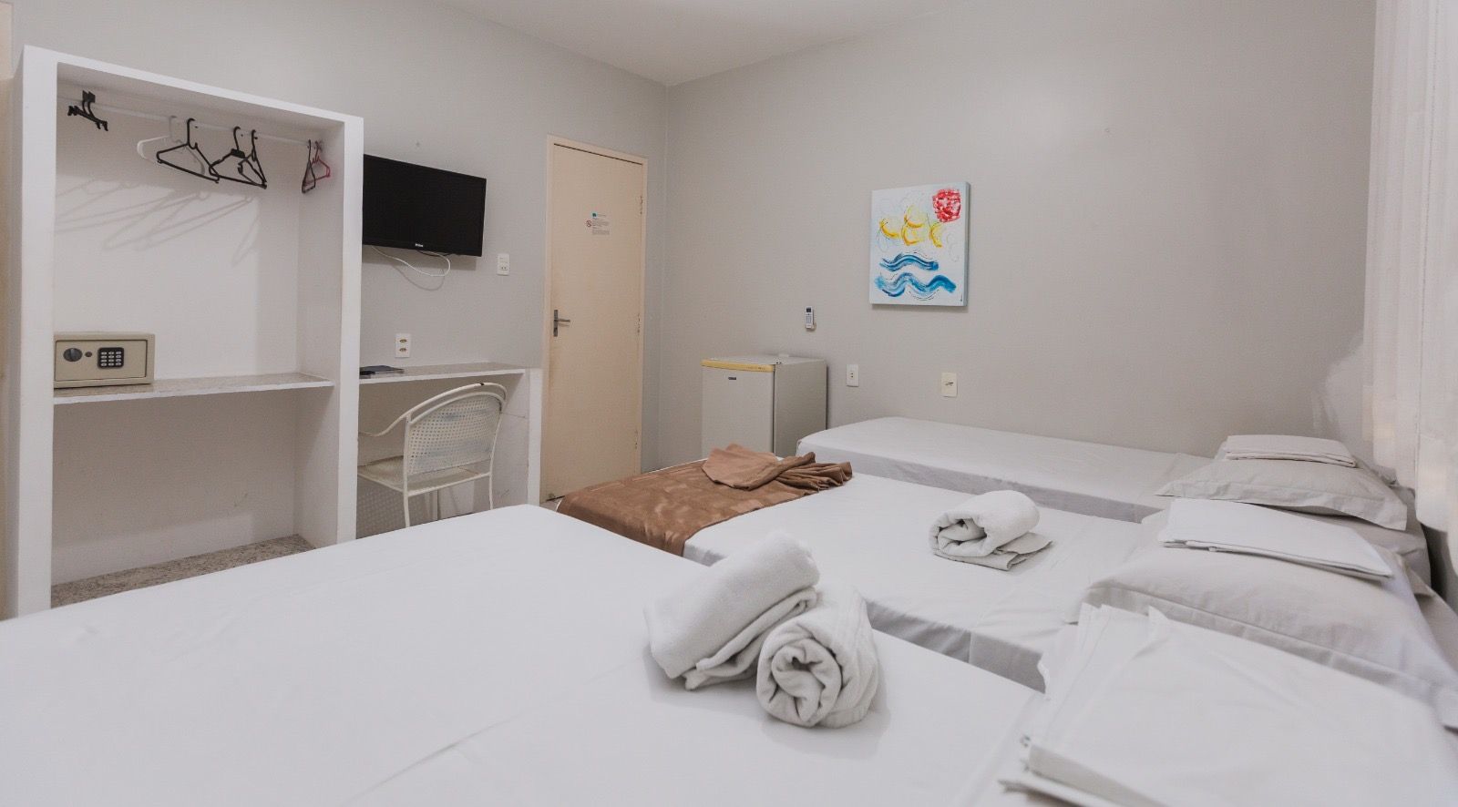 Um quarto de hotel com três camas de lençóis brancos, um armário aberto branco com cofre, uma escrivaninha e obras de arte na parede.