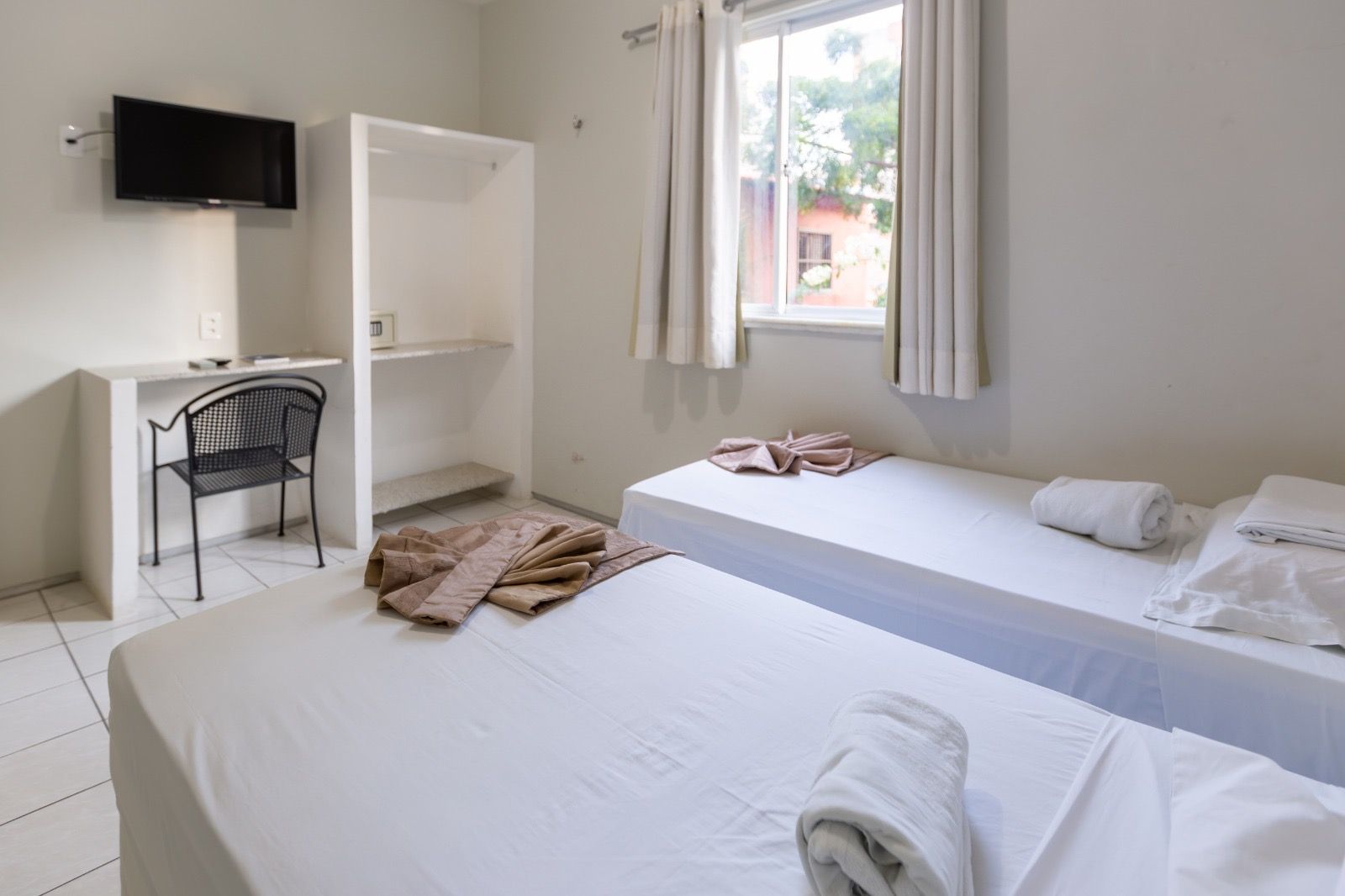 Um quarto de hotel com duas camas de solteiro, roupa de cama branca, uma escrivaninha com uma cadeira preta e uma TV de tela plana fixada na parede.