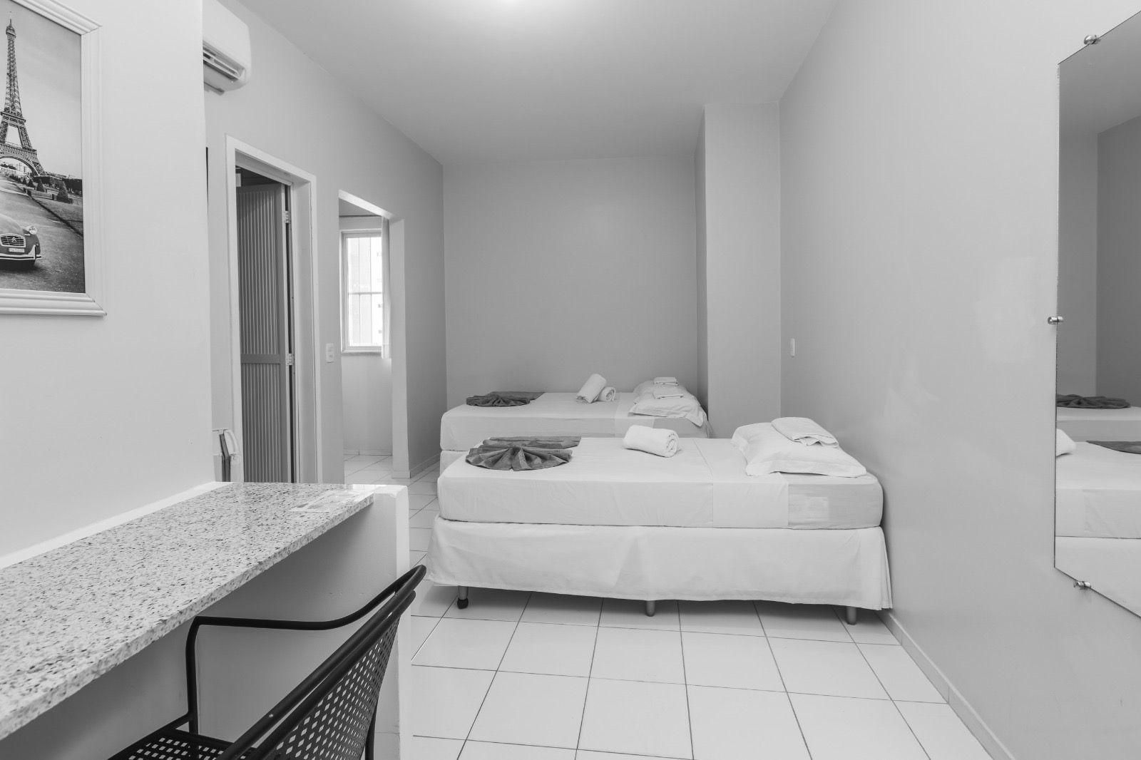 Um quarto de hotel minimalista e bem iluminado, com duas camas de solteiro com lençóis brancos, uma escrivaninha de granito e um espelho de parede.