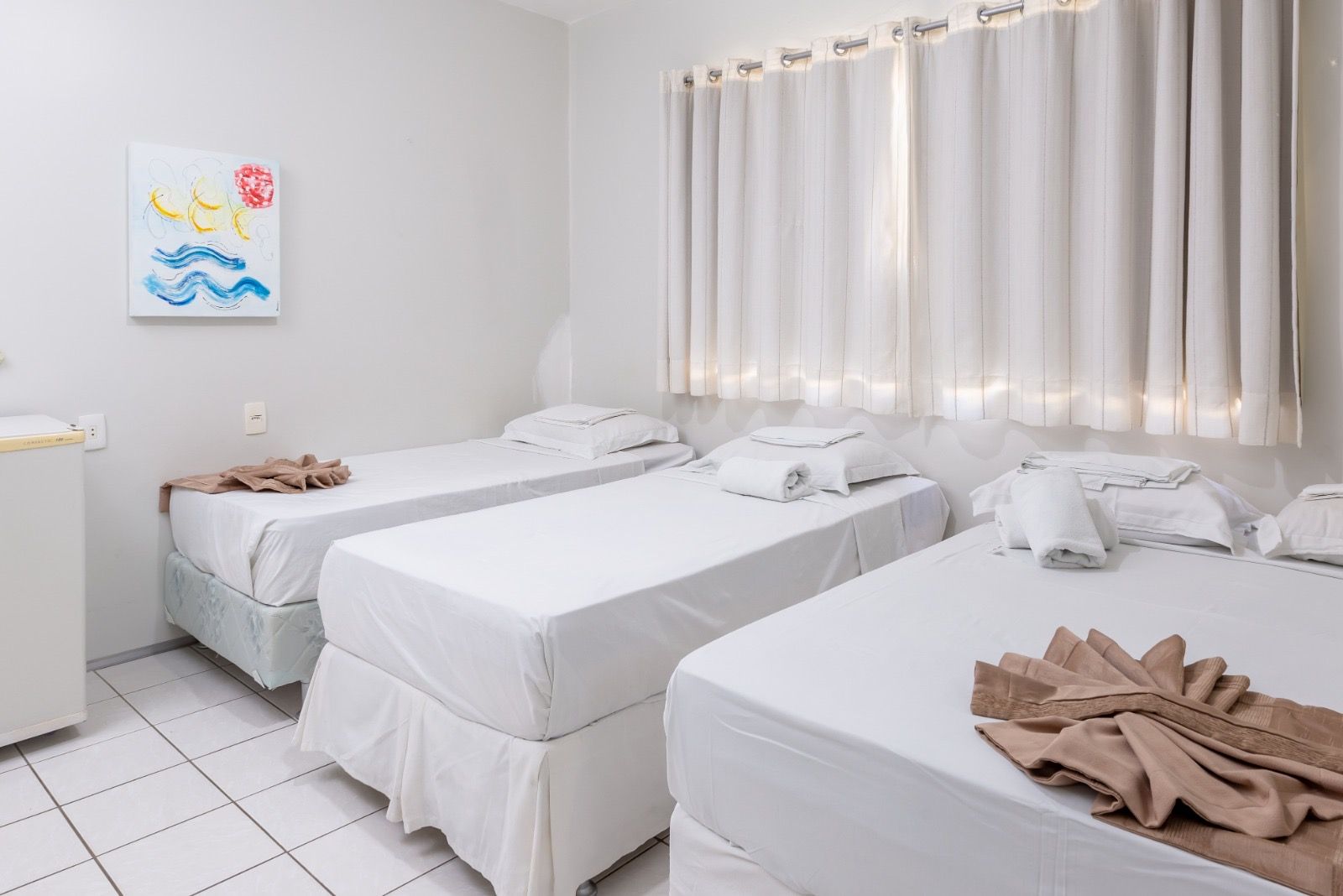 Um quarto de hotel com três camas brancas e limpas, toalhas bege dobradas, piso de azulejo e cortinas brancas na janela.
