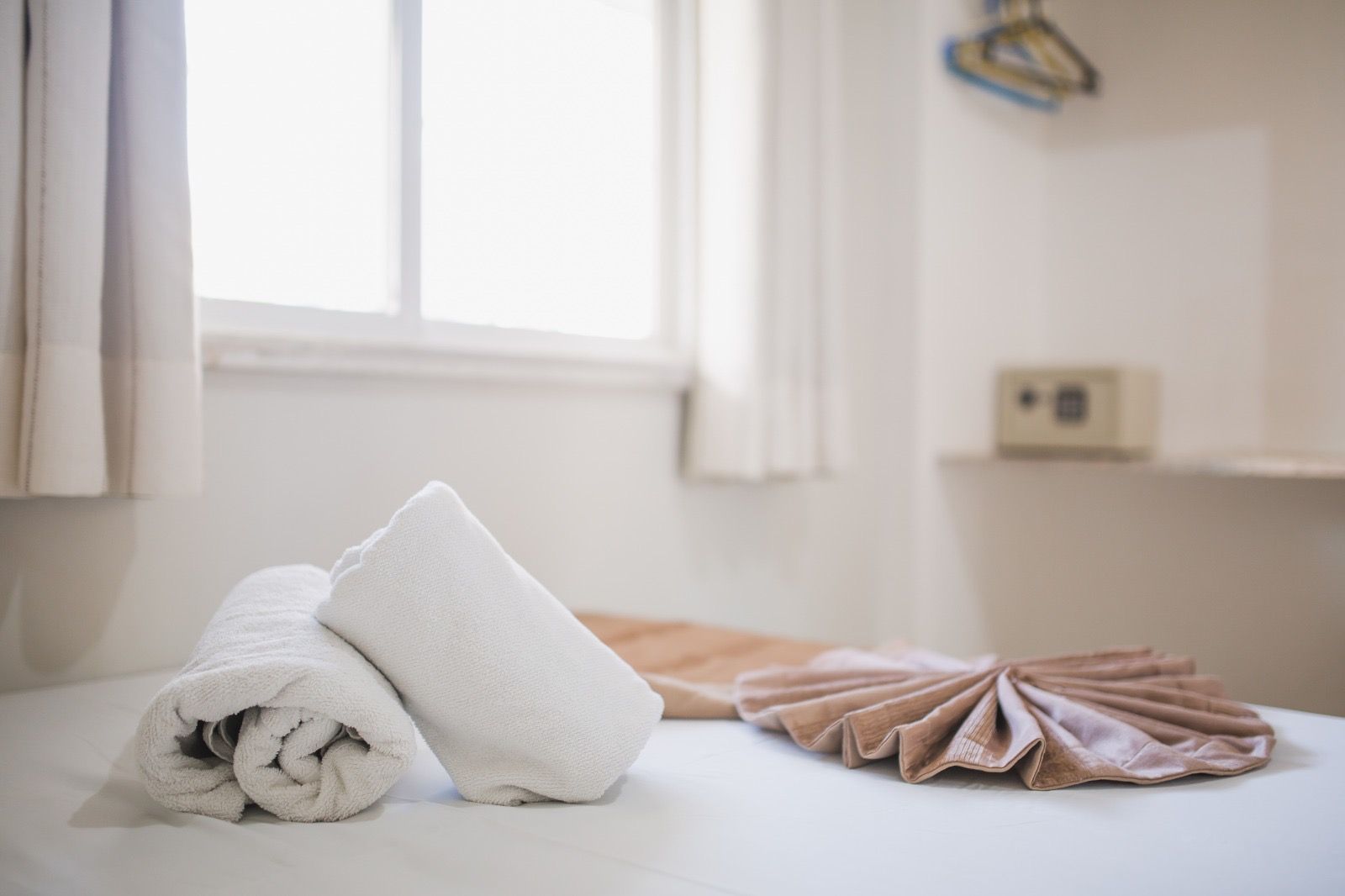 Toalhas brancas enroladas e um leque decorativo de tecido marrom estão sobre uma cama branca em um quarto de hotel, com um cofre embutido na parede ao fundo.