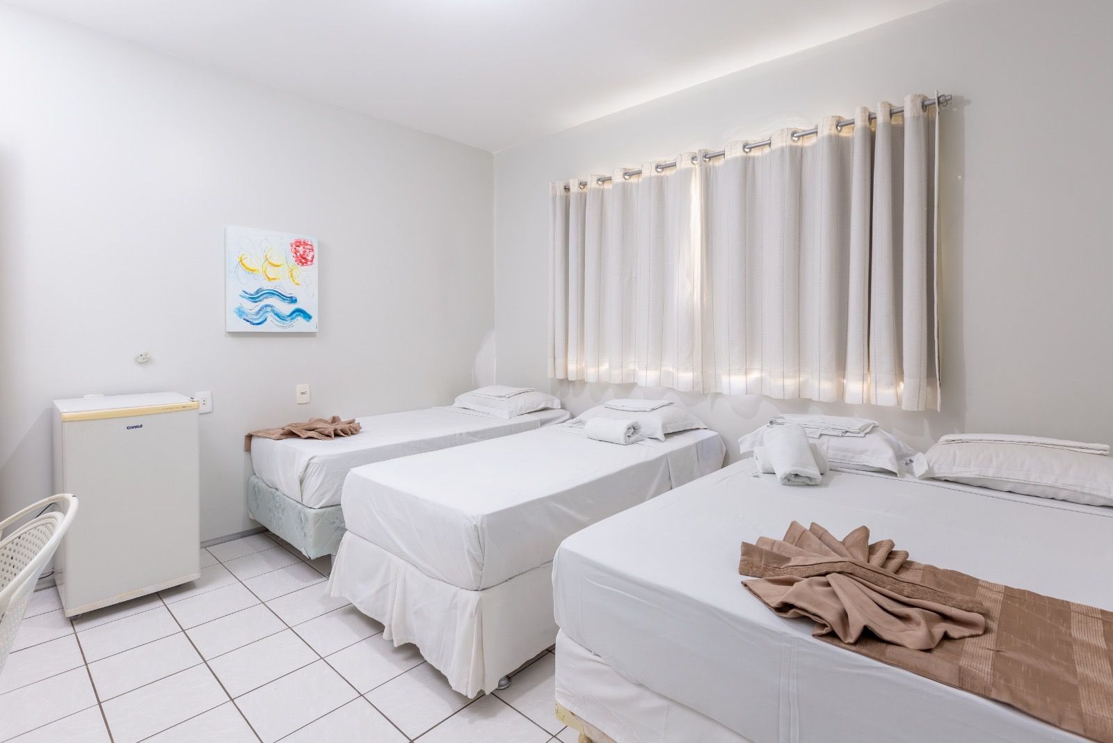 Um quarto de hotel limpo com uma cama de casal e duas camas de solteiro, todas com lençóis brancos, piso de azulejo e frigobar ao lado da cama.