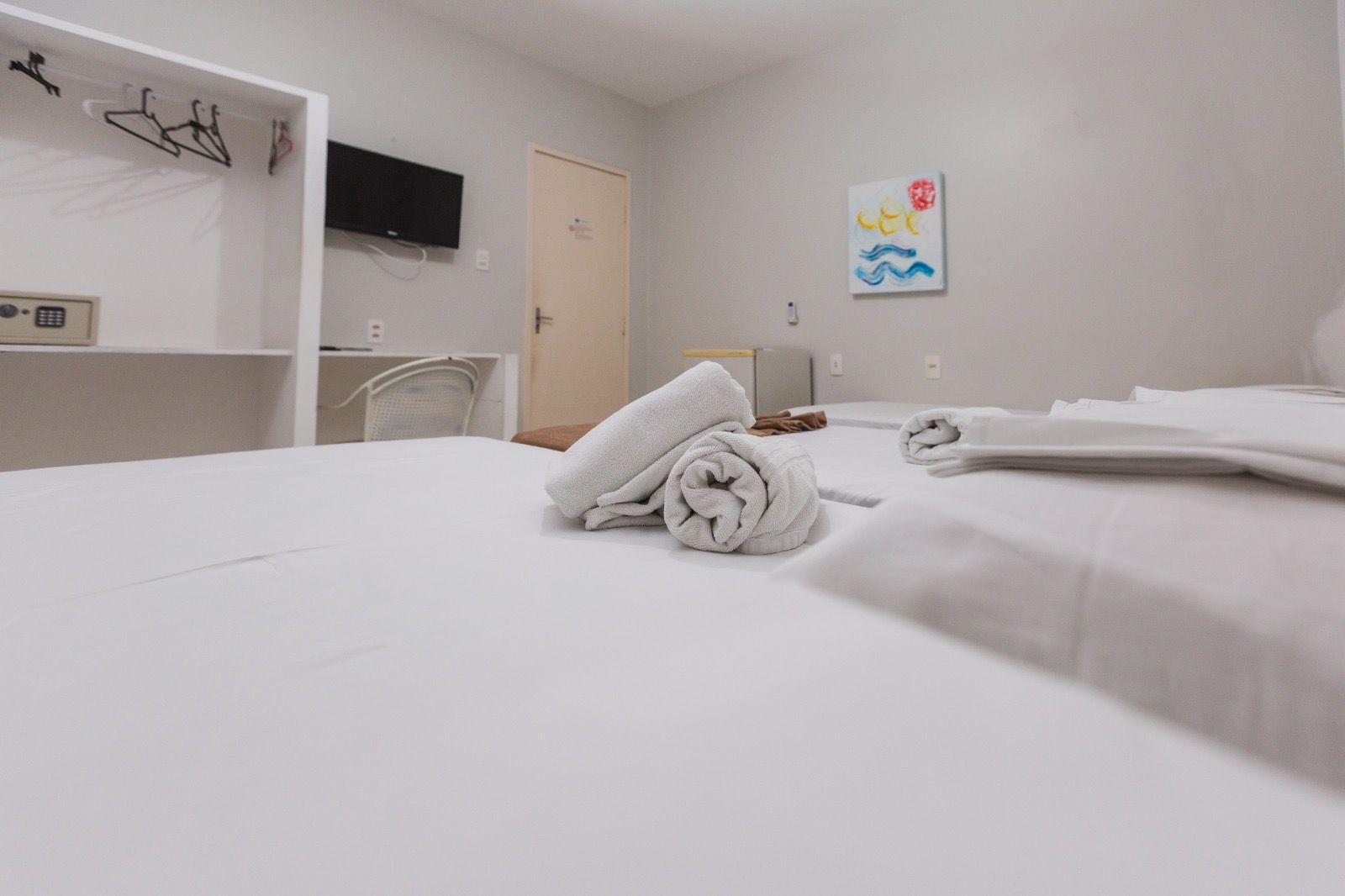 Um quarto de hotel com uma cama branca impecavelmente arrumada, toalhas enroladas no centro, um guarda-roupa aberto e arte minimalista na parede.