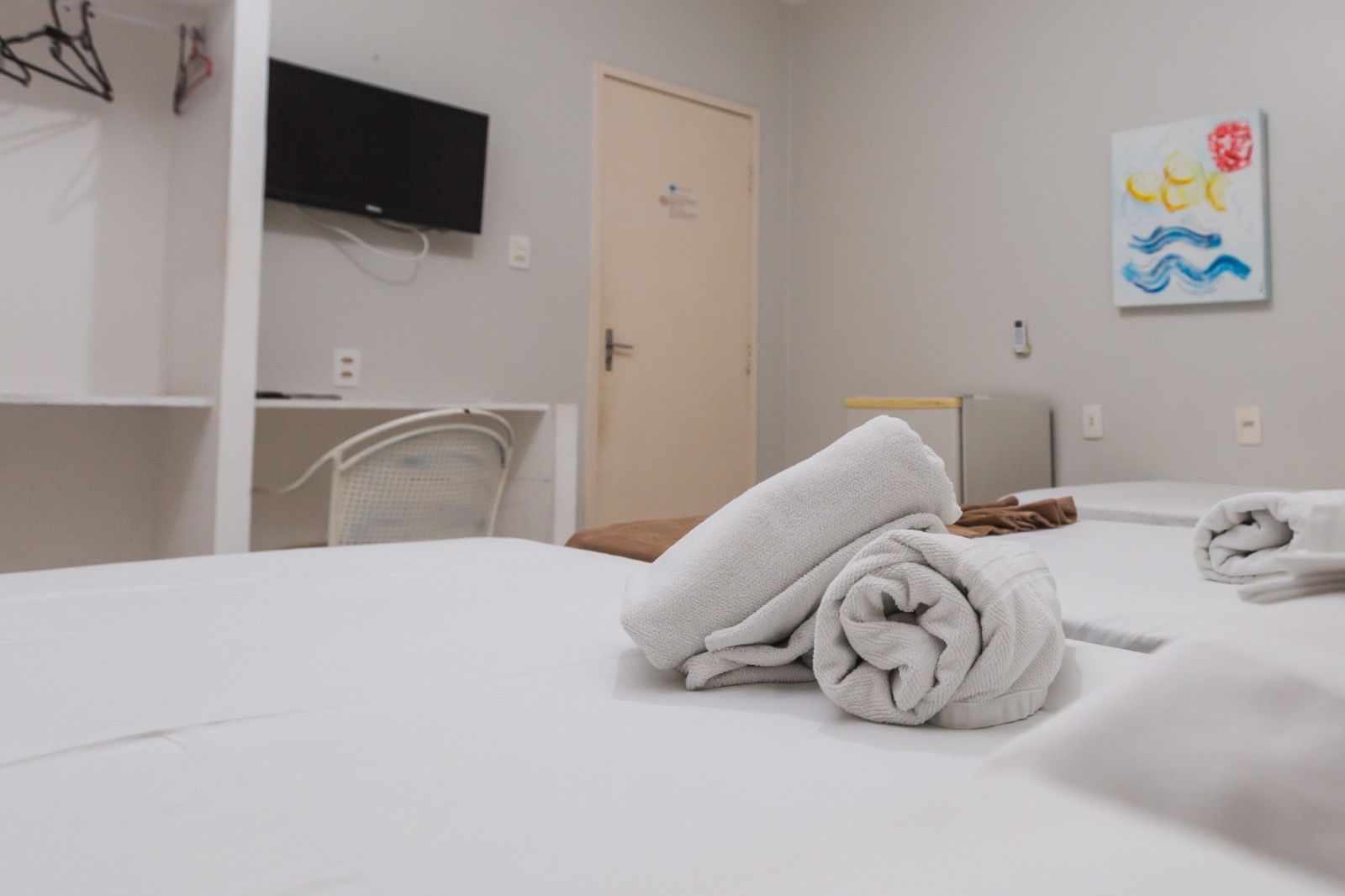 Um quarto de hotel com uma cama bem arrumada, lençóis brancos, toalhas enroladas, uma televisão na parede e uma obra de arte na parede.