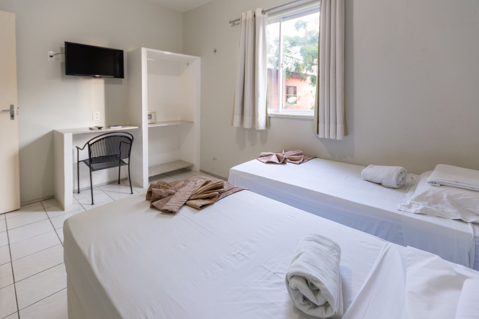 Um quarto de hotel simples com duas camas de solteiro, uma escrivaninha, uma cadeira, um armário aberto e uma TV fixada em uma parede branca lisa.