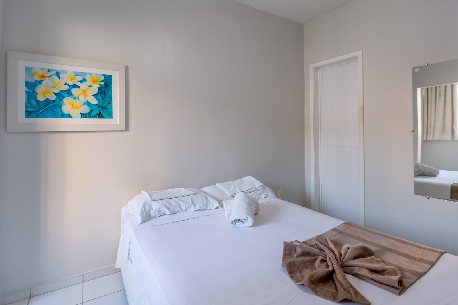 Um quarto de hotel com paredes brancas, uma cama com lençóis brancos, uma manta marrom dobrada e uma pintura floral na parede.