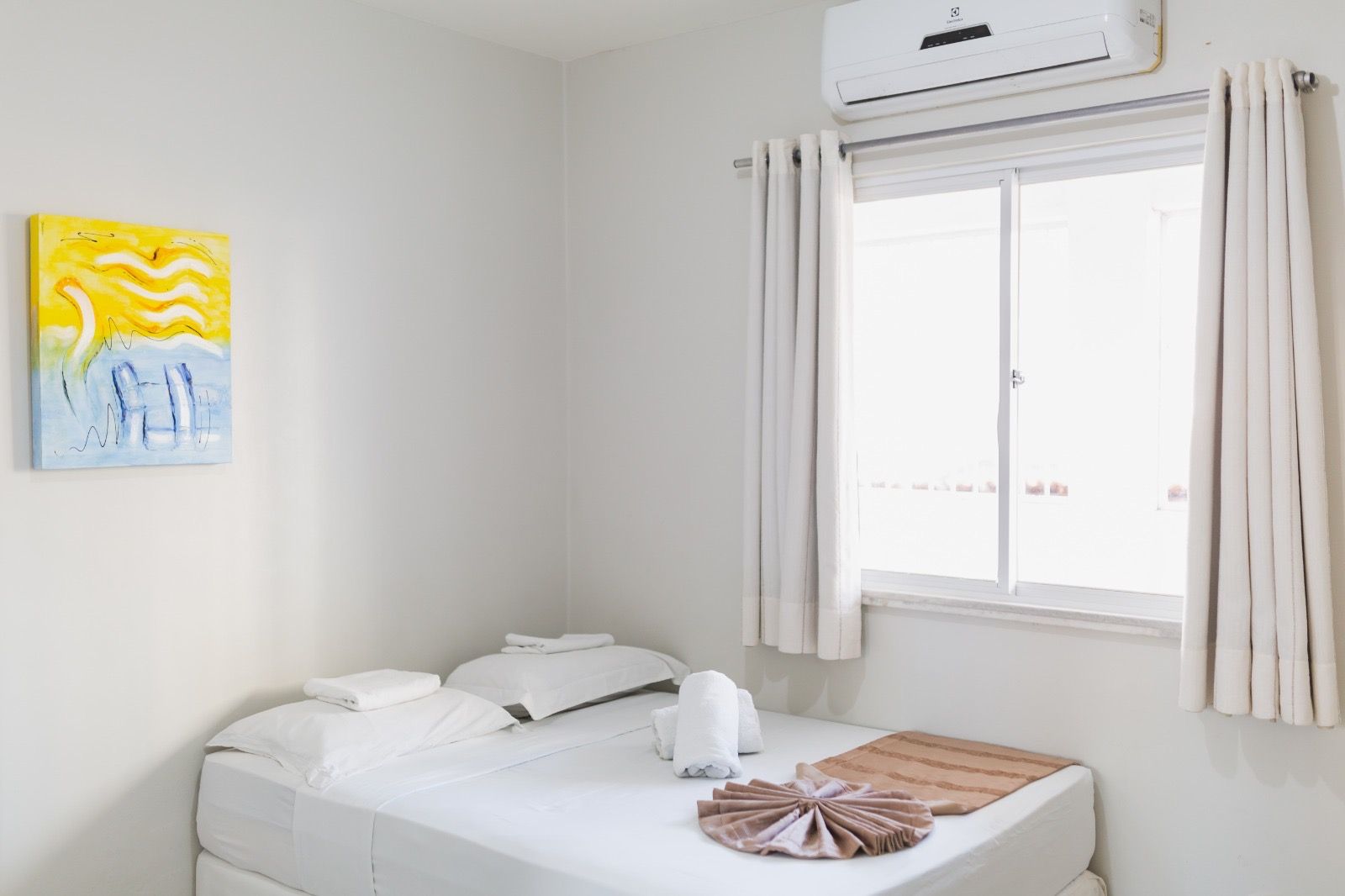 Um quarto com paredes brancas, cama arrumada, dois travesseiros brancos, um suporte para toalhas, uma janela com cortinas brancas e obras de arte na parede.