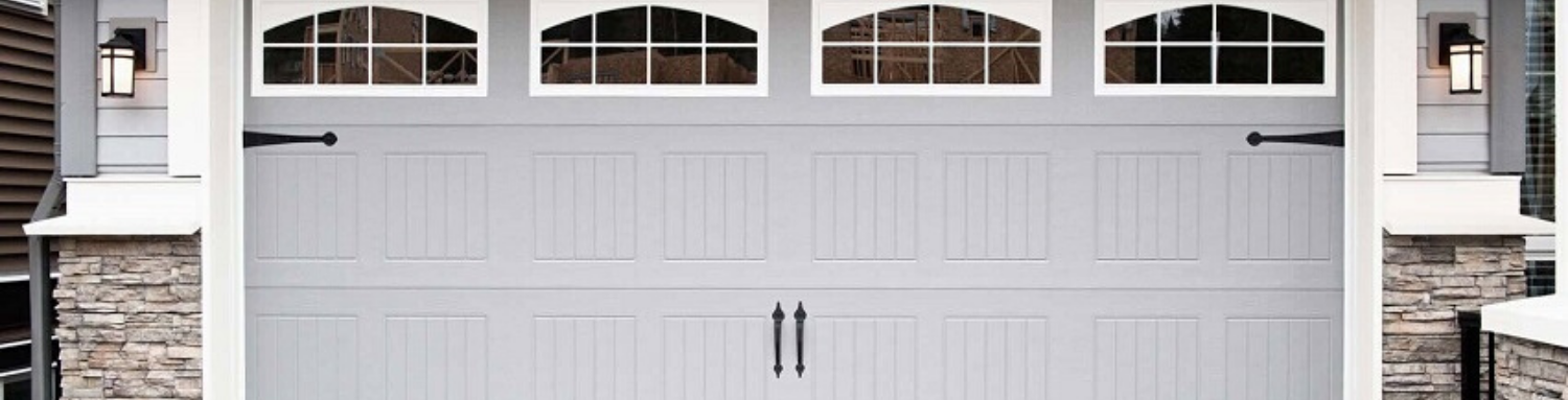 A gray, multi-panel residential garage door featuring decorative black hinges, handles, and arched windows with grid panes.